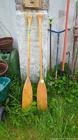Two vintage wooden paddles standing upright against a white wall, showing full length and overall condition.