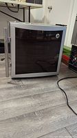Front angle view of compact GE wine chiller with black tinted glass door and silver handle, standing on wood floor.