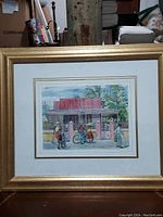Front view of framed watercolor painting depicting a street scene with people and a bicycle in front of a house with a red roof, pink pillars and gate. White matting and gold frame.