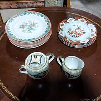 Image showing 9 floral rimmed plates by Grindley & Co., 4 plates with red floral design by Montgomery Ward, and Noritake sugar bowl and creamer on round wood table.