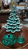 Dark green ceramic Christmas tree with visible layered branches covered in white snow accents and multicolored bulbs, two green ceramic bases placed in front, one larger with winding key