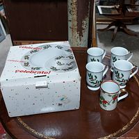 Primary photo showing the china set arranged next to its original box featuring 'celebrate!' branding and holly decorations. Includes 4 plates, 4 saucers, 9 cups total (4 matching cups plus 5 additional holly cups).