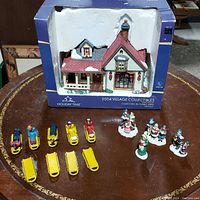 Holiday Time 2004 Village Collectibles farmhouse in original blue box with four ceramic Christmas people figurines and four yellow sleds with sledders arranged in front on a round wooden table.