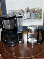 Ninja coffee maker and Culinary Obsession coffee pot placed on a wooden round table with a framed art print in the background.