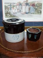 Front view of large brown and white crock and smaller brown flower pot on table with framed artwork behind.