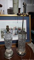 Photo showing two vintage crystal lamps with intricate cut and etched glass designs, metal bases and attached cords, displayed in front of framed artwork.