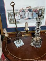 Two vintage crystal lamps on a table in front of a framed painting. One lamp has a marble-like base with three brass arms and no shades, the other is a stacked crystal block lamp with an etched design and a single brass light bulb holder on top. Used condition with cords visible and no bulbs or shades included.