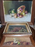 Three fruit-themed paintings shown together: front-facing view of large oil painting by D. Lowe on top with bowl spilling fruit; smaller framed grape painting below; and fruit and cherubs canvas visible at bottom.