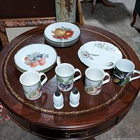 Full view of the stack of ribbed plates, white plates, fruit-themed mugs, single scalloped plate, and salt and pepper shakers on a round wood table.