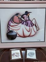 Photo of large pink framed print of two Indigenous women with light pink matting. Below it, two smaller prints in brown wooden frames are visible.