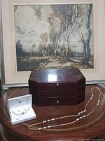 Brown wood octagonal jewelry box with polished finish with lift top and pull out drawer closed, placed in front of a framed artwork. A necklace and earrings set presented in white box next to the box. A long faux pearl detailed necklace laid in front.