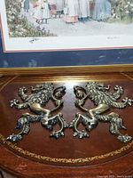 Photo showing both metal lion plaques side by side on a wooden surface beneath a framed picture.
