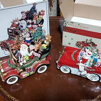Two Fitz and Floyd ceramic Christmas-themed music box cars displayed side by side on a table, one with Santa and reindeer, the other with a snowman holding a list