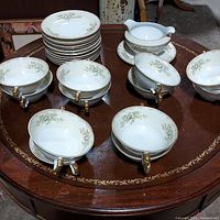 Set of porcelain china items displayed on round wooden table, showing soup bowls, plates, and round server with attached gravy boat and base plate. Floral pattern with gold trim visible on all pieces.