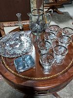 Glass pitcher with floral silver etching, six crystal glasses with silver rims, glass plate with floral silver design, silver lidded dish, glass bell with a silver handle, small glass vase with silver decoration on wooden table.