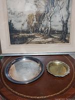 Photo showing both the large round engraved silver platter and smaller round scalloped edge platter displayed on a wooden surface beneath a framed picture.