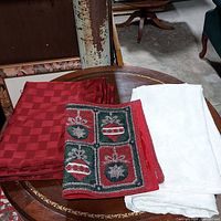 Photo showing folded burgundy tablecloth with woven square pattern, folded ivory tablecloth with subtle floral design, and a Christmas-themed placemat featuring green and red ornament blocks with gold accents, placed together on a wooden oval table.