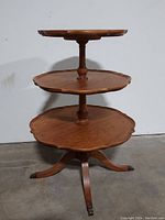Side view of three-tier wooden butler's table showing scalloped edges and turned center supports ending on metal claw feet.