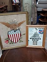 Framed American flag needlepoint and framed WWII coin collection displayed side by side on a surface.