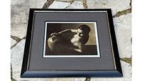 Framed sepia-toned reproduction photograph after Ha Von Behr titled Rebecca 1938 showing a nude woman reclining with a vase, displayed outdoors on pavement.