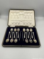 Opened fitted case displaying 12 sterling silver teaspoons and sugar tongs on blue velvet lining