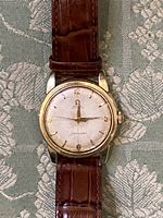 Front view of watch showing textured silver-white dial, applied gold indices, Omega logo, “Automatic” and “Seamaster” text, brown leather strap on patterned fabric