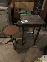 Both small wooden side tables on basement floor showing size and condition: octagonal tripod pedestal side table with reddish worn top and rectangular table with shelf and dark finish.