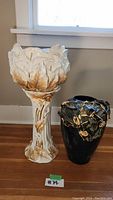 Full front view of white ceramic plant stand with gold leaf detailing and black ceramic vase with gold accents side-by-side on floor.