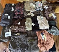 Photo showing multiple tops folded and arranged on table, predominantly earth tones and greys, with visible price tags obscured, indicating new condition and assorted sizes.