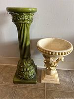 Side view of tall green ceramic plant stand and shorter beige decorative plant stand on floor.
