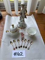 Entire lot displayed on white paper includes the porcelain sugar bowl, teacup, angel figurine, butterfly figurine, and seven decorative teaspoons lined up in front.