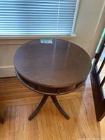 Round wooden side table viewed from an angle showing the drawer and tripod base.