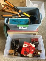Two plastic bins stacked; top bin contains assorted garden tools including shears and saw, boxed item; bottom bin contains assorted smaller tools and hardware items including ear protection box