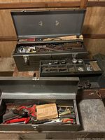 Photo showing two metal toolboxes filled with various hand tools and hardware items, some tools have red handles and there are visible clamps, wrenches, and screwdrivers.