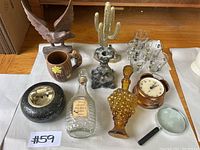 Photo showing vintage home decor items including brass cactus, eagle sculpture, glasses, clock, decanters, ashtray, mug, and magnifying glass on white cloth.
