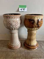 Two brown and beige glazed ceramic plant stands with floral relief designs and fluted pedestal bases shown side by side on tile floor against white wall.