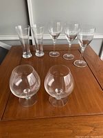 Wide shot of 2 large brandy glasses, 3 wine glasses, and 2 small beer glasses arranged on wooden table.