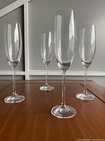 Four clear champagne flute glasses arranged on a wooden table with a white paneled background, showing tall stems and narrow bowls