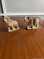 Front and side view of two brown rapoko wood hand carved elephant figurines placed on a wooden surface, showing detailed texture and base dimensions.