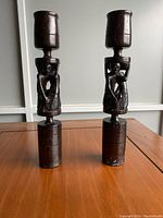 Pair of dark stained hand carved wooden candlesticks with intricate abstract designs carved into the middle section, set on a wooden surface, showing a rich wood grain and wax residue.