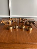 Front view of 4 hand carved wooden napkin holders shaped as 2 elephants, 1 lion, 1 zebra, and 4 gold-tone ribbed metal cylindrical napkin rings on wooden surface