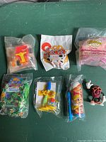 Seven vintage small toys still sealed in their original plastic packaging, laid out on a green surface, showing various characters and themes from early 1990s including McDonald's Happy Meal toys and cereal prizes.