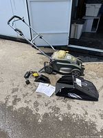 Photo of the full lawn mower with foldable handle, grass collection bag, battery charger, discharge chute and manuals laid out on ground.