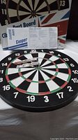 Front side of the Cooper dart board with 5 brass darts and dart board rules sheets behind it