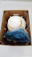 Top-down view of cardboard box containing white and beige plates stacked together with blue plastic container on top containing utensils.