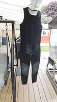 Front view of black neoprene scuba pants hanging on a hanger outside on deck. Pants have reinforced knee pads and visible stitching.