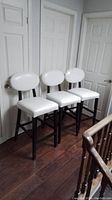 Set of three white bar stools with round backrests and black wooden frames, standing on a wooden floor near white doors
