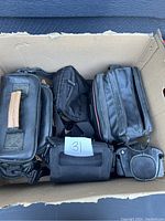 Top-down view of six assorted black camera bags inside a cardboard box, showing different styles and sizes, one has a wooden handle.