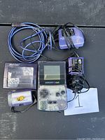 Photo displays a translucent purple Gameboy Color handheld console with visible buttons and screen, accompanied by two additional battery packs, a Mad Catz battery pack, and a blue AV cable for video output.