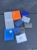 Ten assorted plastic Gameboy dust protector cases in various colors and sizes laid out on a dark surface with a handwritten label showing number 39.